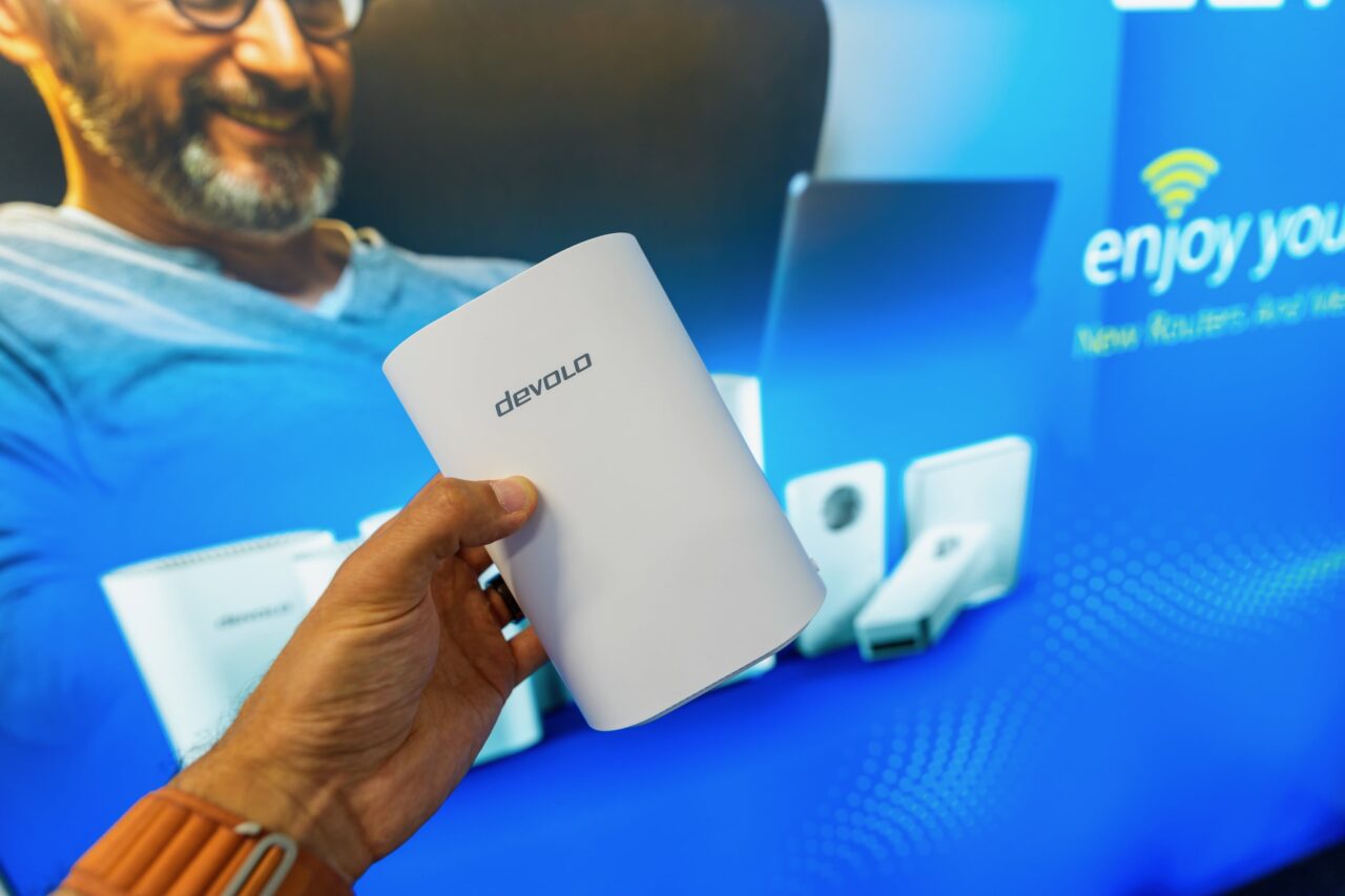 devolo BE6500 Boîtier Wi-Fi devolo blanc tenu en main devant le panneau du stand.