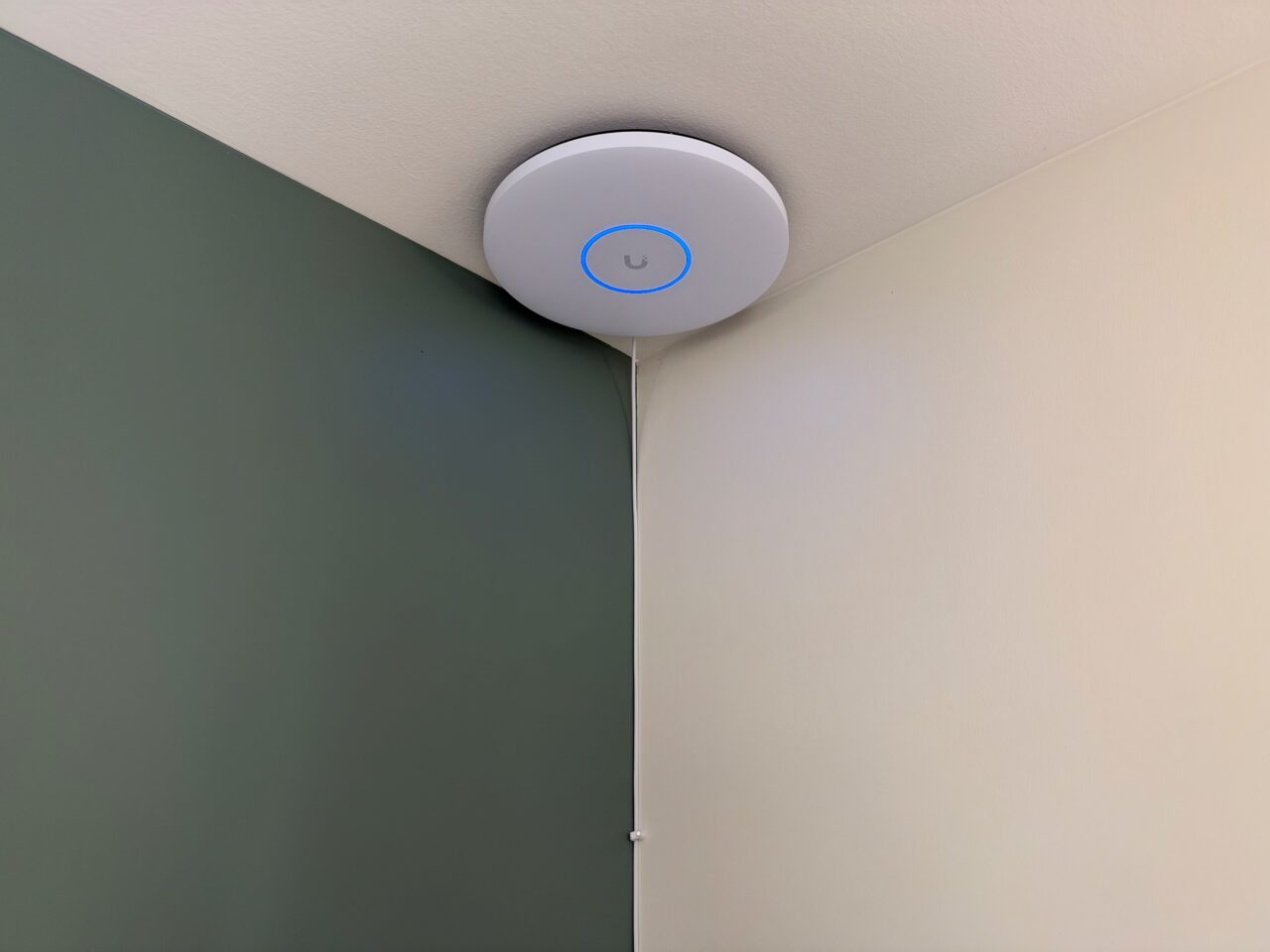 Borne UniFi U7 Pro XGS allumée au plafond, avec son câble Ethernet blanc descendant dans l'angle des murs.