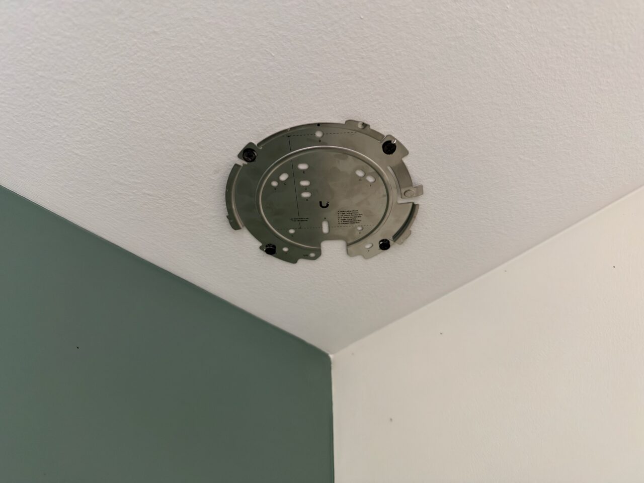 Support de la borne UniFi U7 Pro XGS fixé au plafond en placo par quatre vis, près du coin de la pièce.